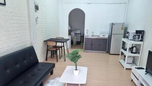 Een knus woongedeelte met een zwarte bank, een witte tafeltje met een plant, een kitchenette en een televisie op een standaard.