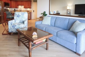 A living room with a sofa, a coffee table, a chair, and a kitchen area in the background. - Kihei Ocean View Haven | 1BR • AC • Pool • Kitchen (Kihei)