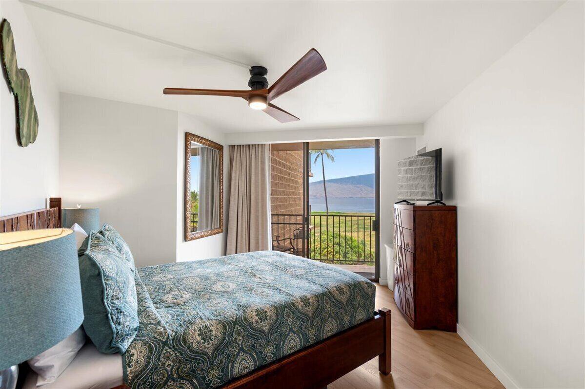 A bedroom with a bed, a ceiling fan, a nightstand, a dresser, and a balcony with a view of the beach and ocean.