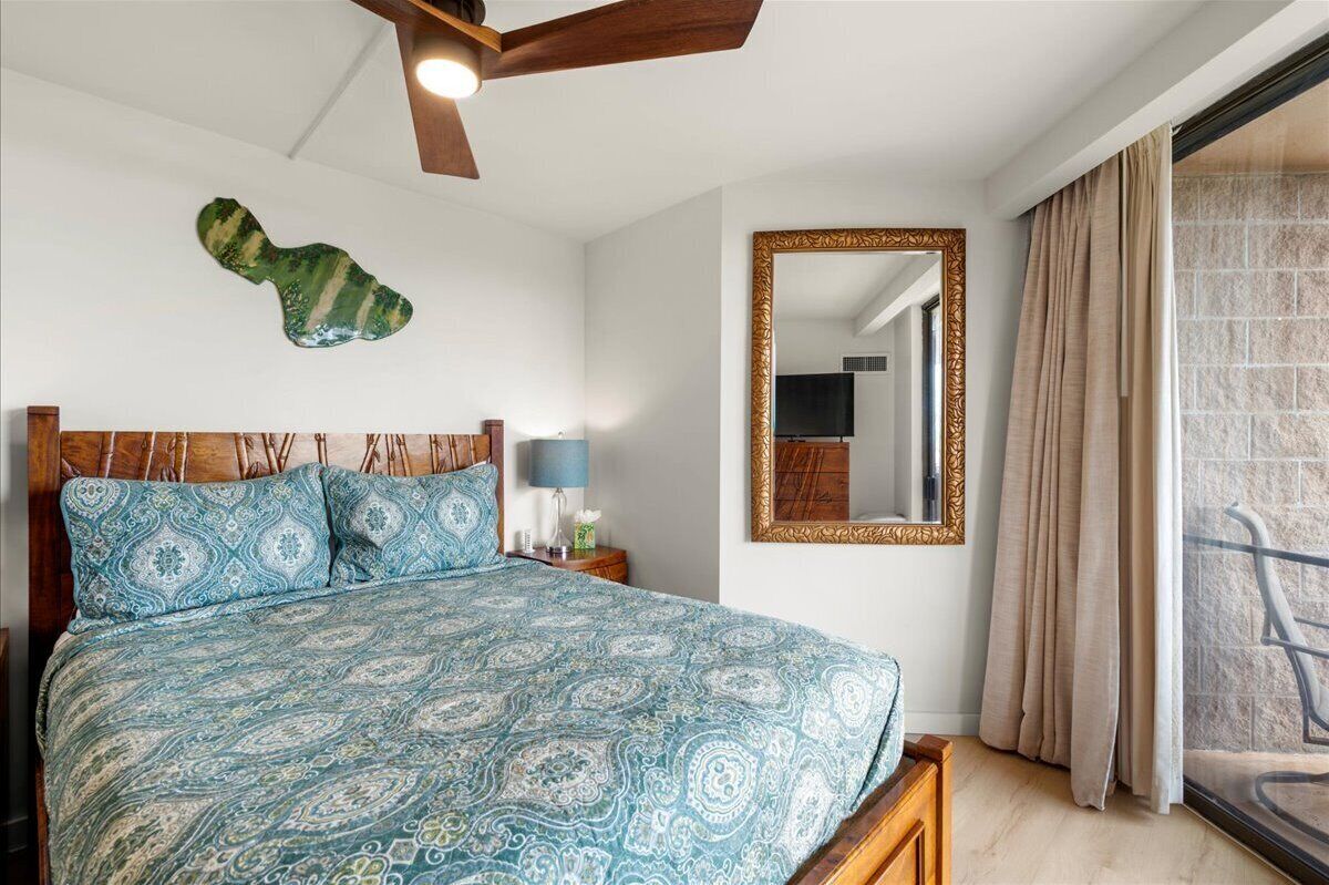 A bedroom with a patterned bedspread, a wooden headboard, a ceiling fan, a large mirror, and a view of the outside through a window.