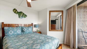 A bedroom with a patterned bedspread, a wooden headboard, a ceiling fan, a large mirror, and a view of the outside through a window.
