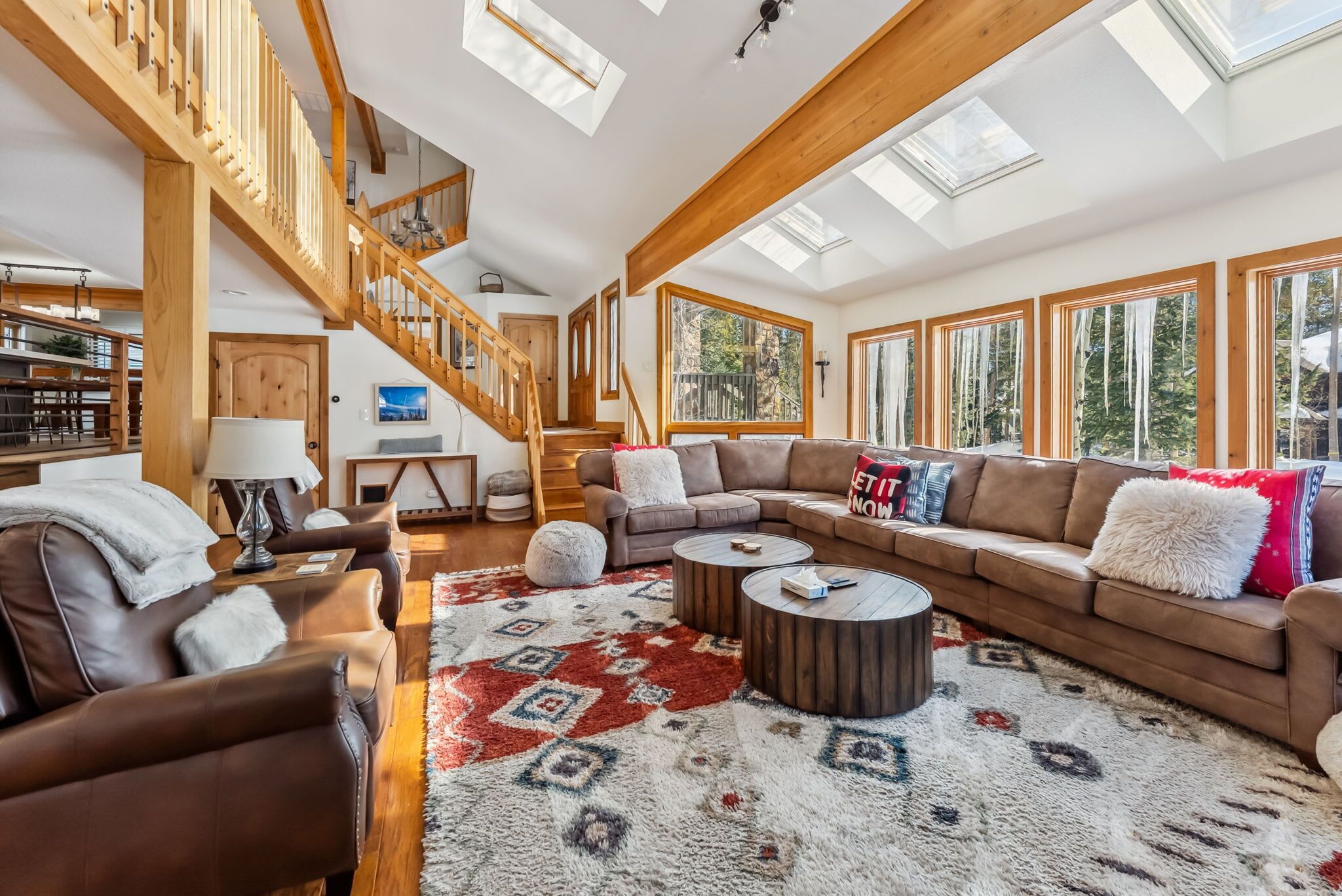 Ein geräumiges Wohnzimmer mit hoher Decke, Holzbalken, einer Treppe und großen Fenstern mit Blick ins Freie.