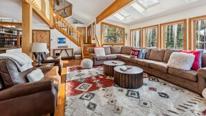 Amplia sala de estar con techo alto, vigas de madera, una escalera y grandes ventanas que ofrecen vistas al exterior.