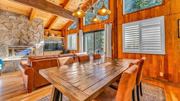 A spacious wooden dining area with a large table and leather seating, a stone fireplace, and large windows with shutters.