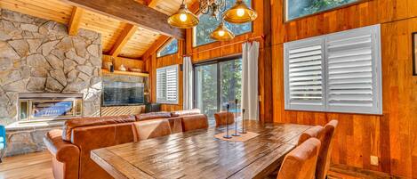 A spacious wooden dining area with a large table and leather seating, a stone fireplace, and large windows with shutters.