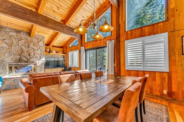 A spacious wooden dining area with a large table and leather seating, a stone fireplace, and large windows with shutters.