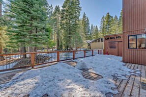 A wooden deck with snow, a metal fence, and tall evergreen trees.
