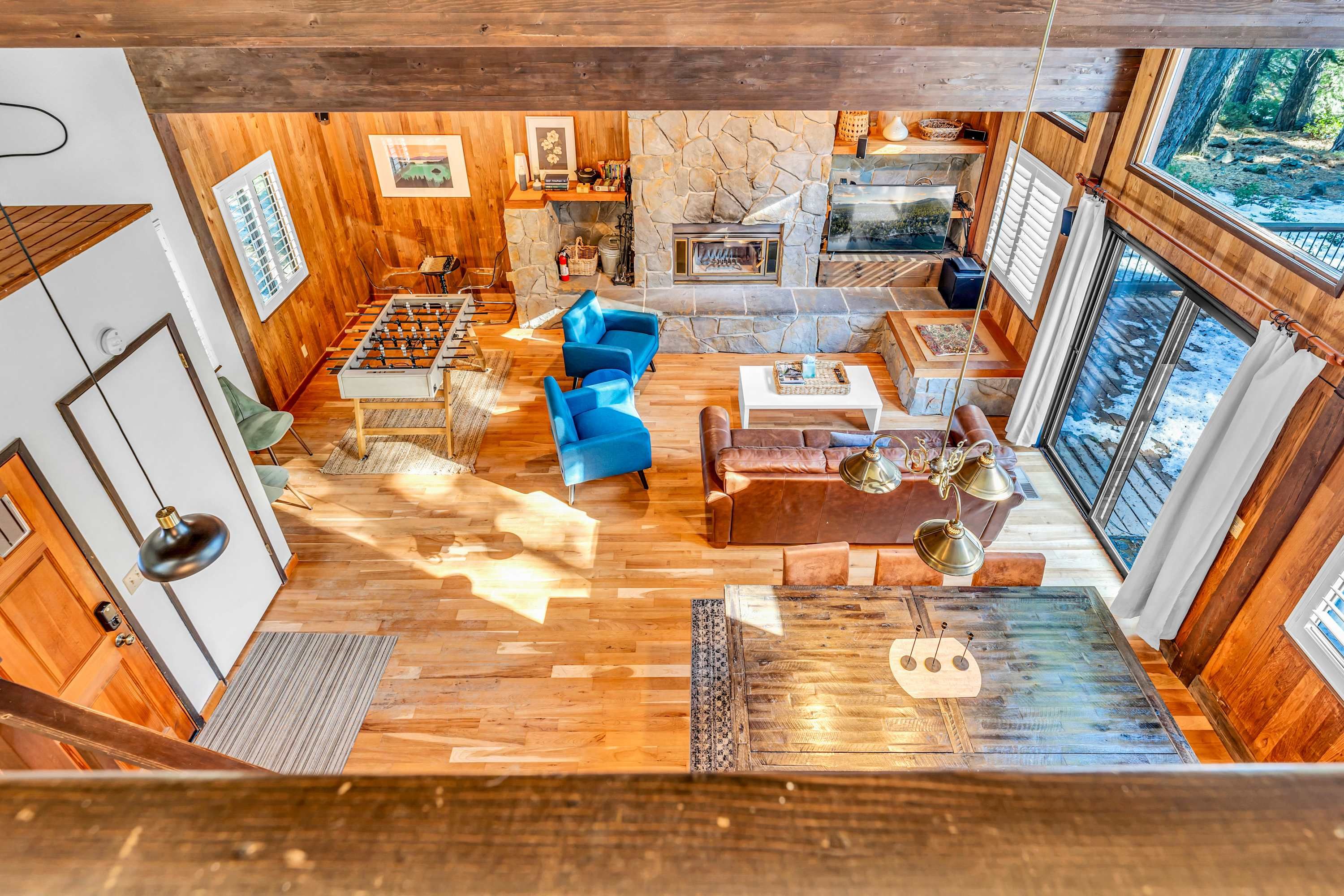 A spacious living area with a stone fireplace, wooden floors, blue sofas, and a large window overlooking a forest.