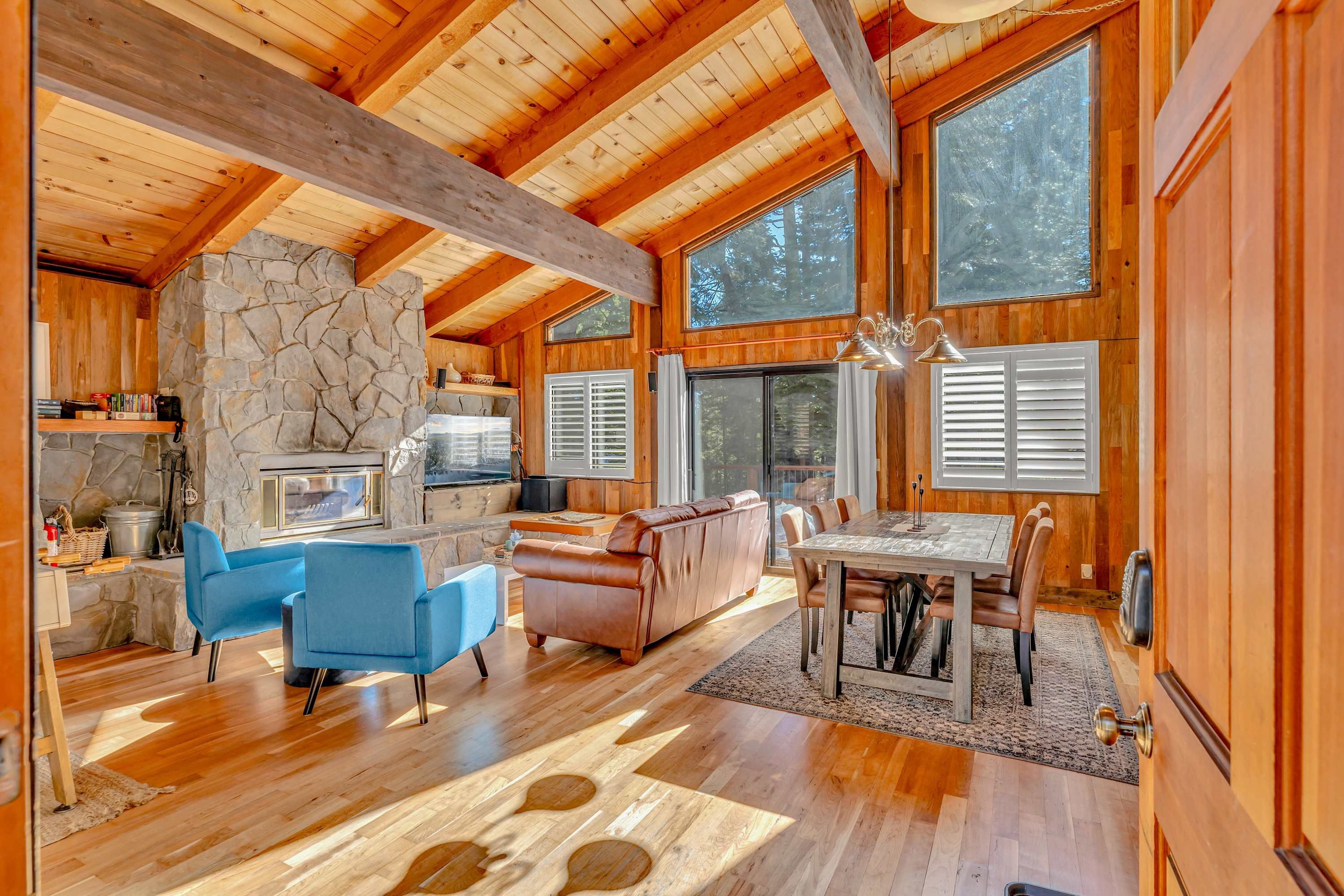 A spacious cabin with a stone fireplace, wooden ceiling beams, and large windows offering views of the forest.
