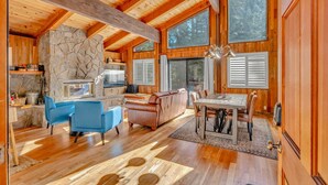 A spacious cabin with a stone fireplace, wooden ceiling beams, and large windows offering views of the forest.