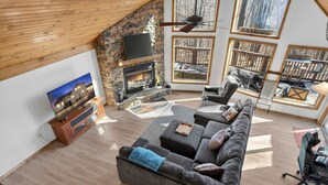 A spacious living room with a stone fireplace, a large sofa, and multiple windows offering views of the outdoors. - Cabin w/hot tub, fire pit & deck (Hedgesville)
