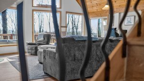 A modern living room with a grey sectional sofa, a wooden ceiling, and large windows offering a view of bare trees. - Cabin w/hot tub, fire pit & deck (Hedgesville)