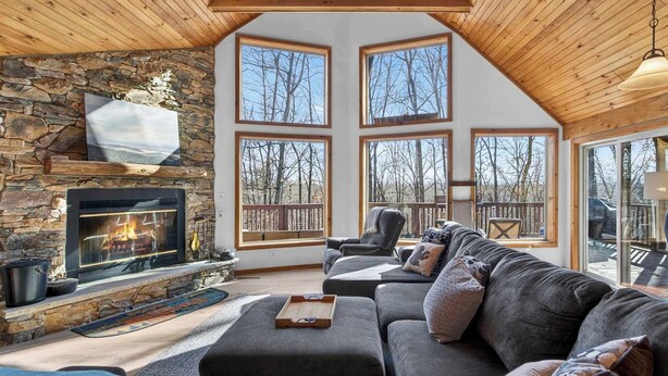 A cozy living room with a stone fireplace, large windows, a wooden ceiling, and a comfortable seating area. - Cabin w/hot tub, fire pit & deck (Hedgesville)
