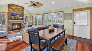 A spacious living room with a stone fireplace, large windows, and a dining area with a wooden table and benches. - Cabin w/hot tub, fire pit & deck (Hedgesville)