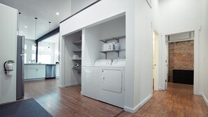A laundry room with a washing machine and dryer, a brick wall, and a kitchen area in the background.