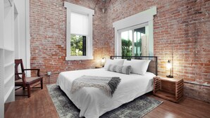 A bedroom with a brick wall, a large bed, two bedside tables with lamps, a chair, and a window.