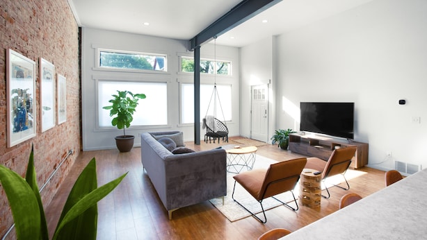 A modern living room with a grey sofa, brown chairs, a TV, and a brick wall.