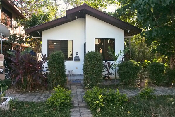 Una casa moderna de dos pisos con fachada blanca y techo marrón, rodeada de vegetación exuberante y un camino pavimentado que lleva a la entrada.