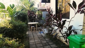 A paved pathway leading to a small outdoor seating area with a table and chairs, surrounded by lush greenery and a green trash bin. - Casa Rafael Coron (Coron)