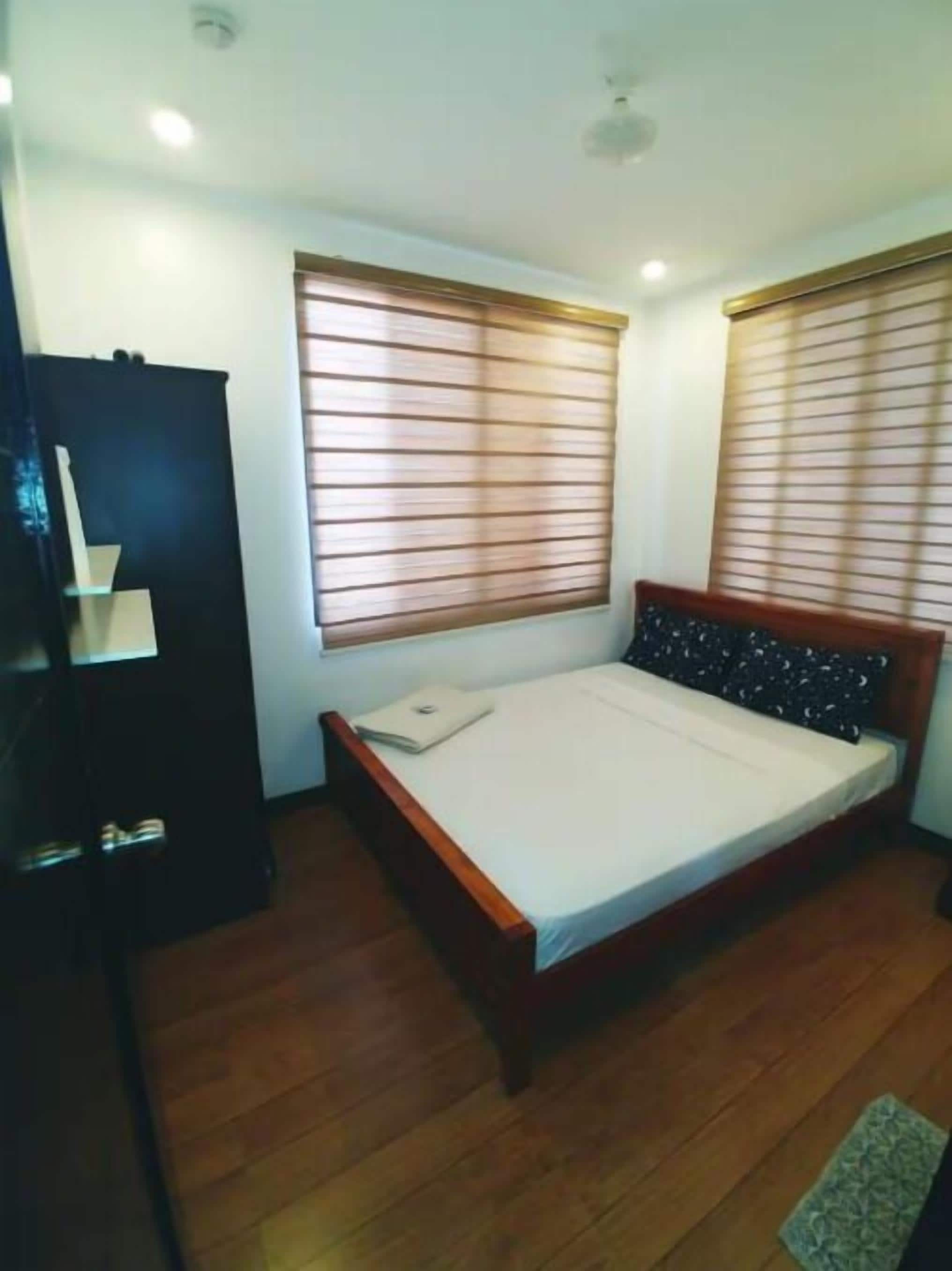 Une chambre à coucher avec un lit, deux fenêtres équipées de stores, une armoire et un petit tapis.