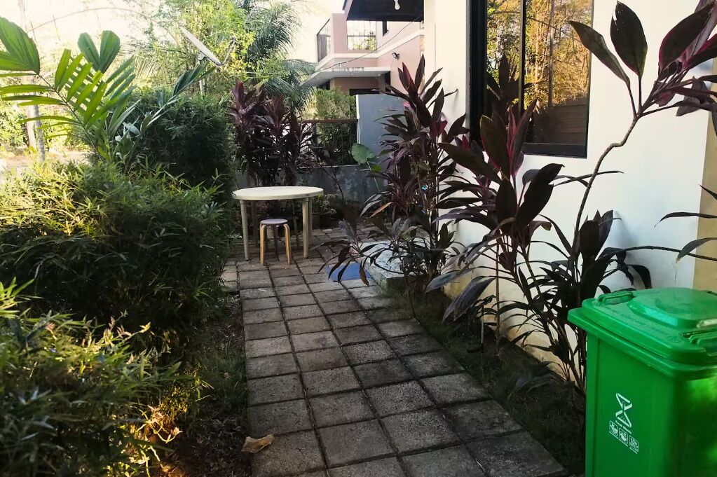 Un chemin pavé mène à un petit espace extérieur aménagé avec une table et des chaises, entouré d’une végétation luxuriante et agrémenté d’une poubelle verte.