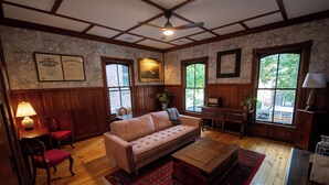 Un salon chaleureux avec un canapé, des fauteuils, une table basse et une cheminée. Des certificats encadrés sont accrochés au mur, et une grande fenêtre offre une vue sur les arbres.