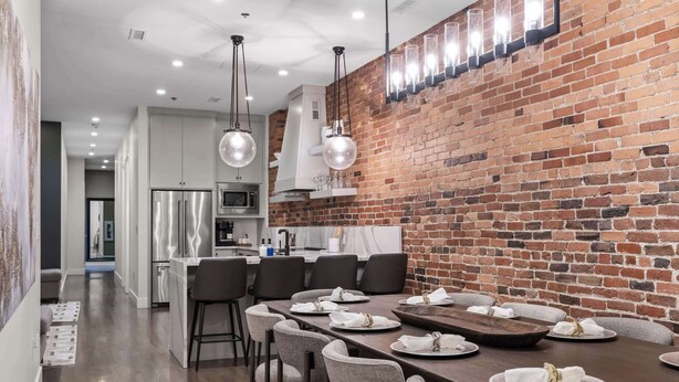 A modern kitchen with a brick wall, stainless steel appliances, and a dining area with a long table set for six. - French’s Loft | Luxe Stay Near Broadway (Nashville)
