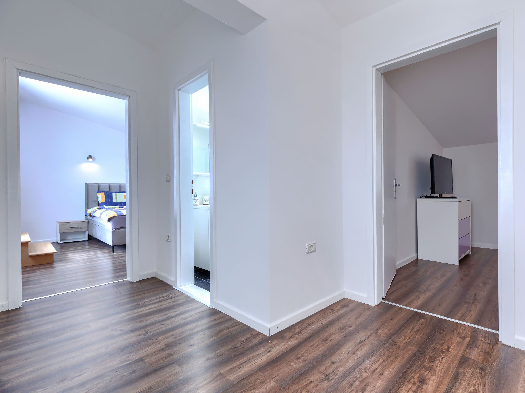 Eine moderne Wohnung mit Holzdielenböden, weißen Wänden, einem sichtbaren Schlafzimmer mit Bett und Nachttisch sowie einem kleinen Fernseher auf einem Schrank.
