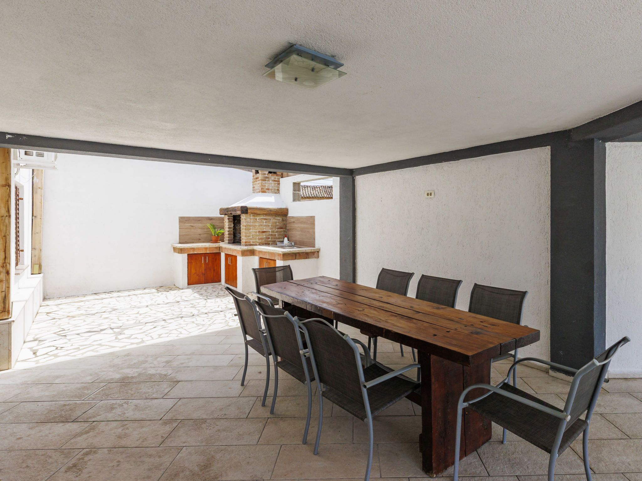 A dining area with a wooden table and chairs, a brick oven, and a small kitchen area with cabinets and a potted plant.