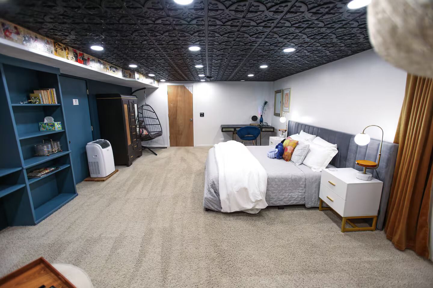 A bedroom with a bed, bedside table, lamp, and a desk with a chair. There is a wardrobe and a heater on the floor.