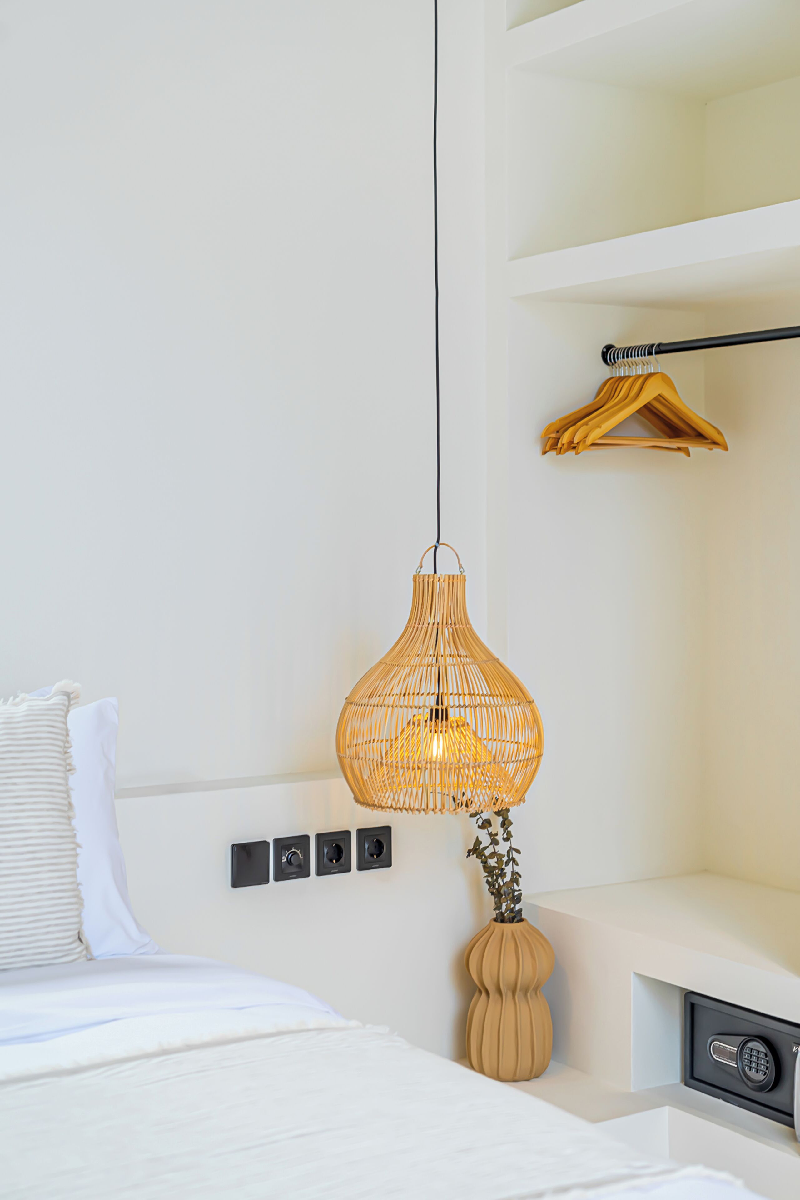 Una habitación de hotel moderna con una cama, una lámpara de mimbre colgante, un perchero de madera y un pequeño jarrón con una planta.