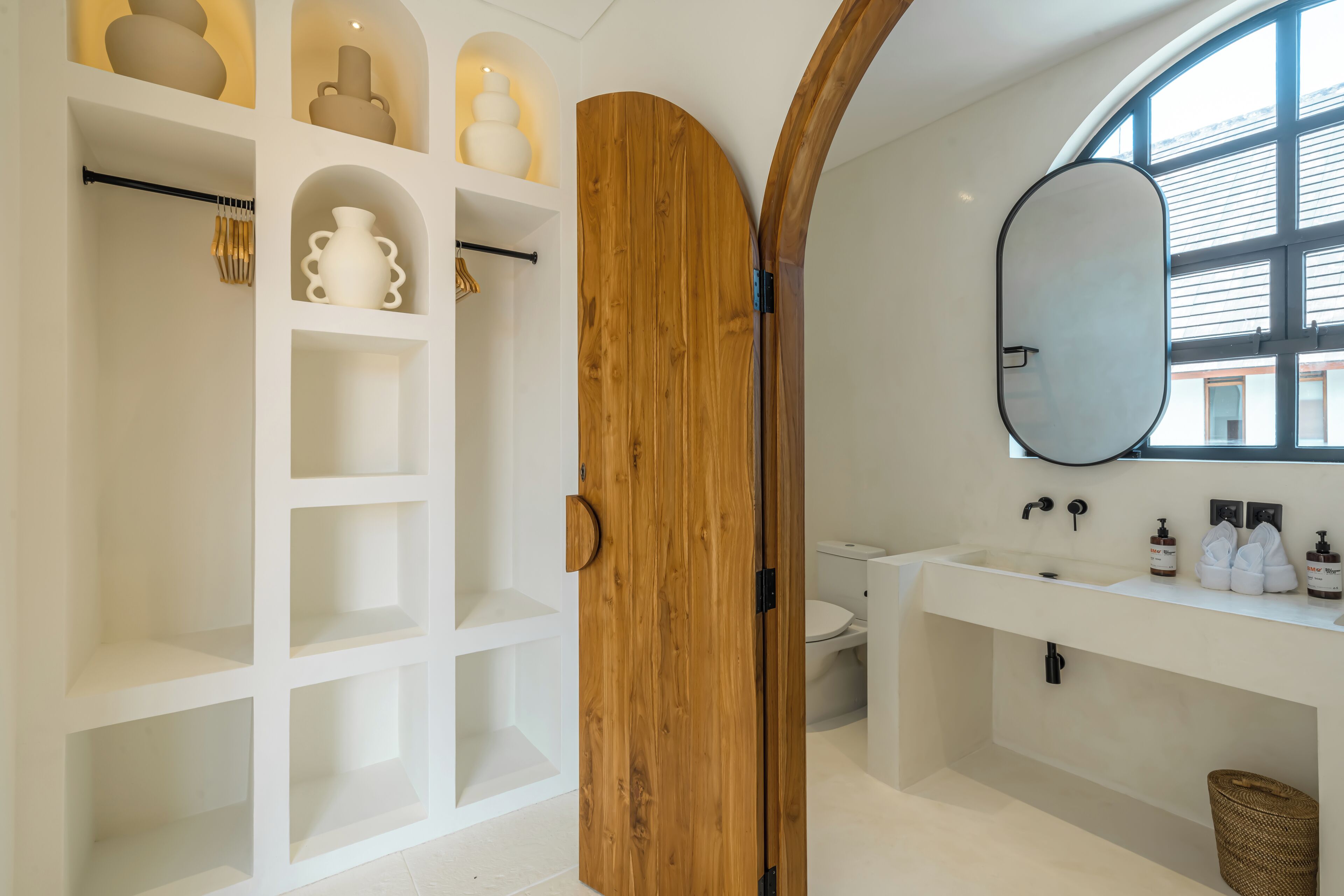 Une salle de bain moderne avec une porte en bois, des étagères blanches, un grand miroir et un lavabo blanc.