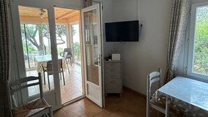 Ein Zimmer mit Esstisch, Stühlen, einem an der Wand befestigten Fernseher und einer Glastür, die zu einem Außenbereich mit Tisch und Stühlen führt.