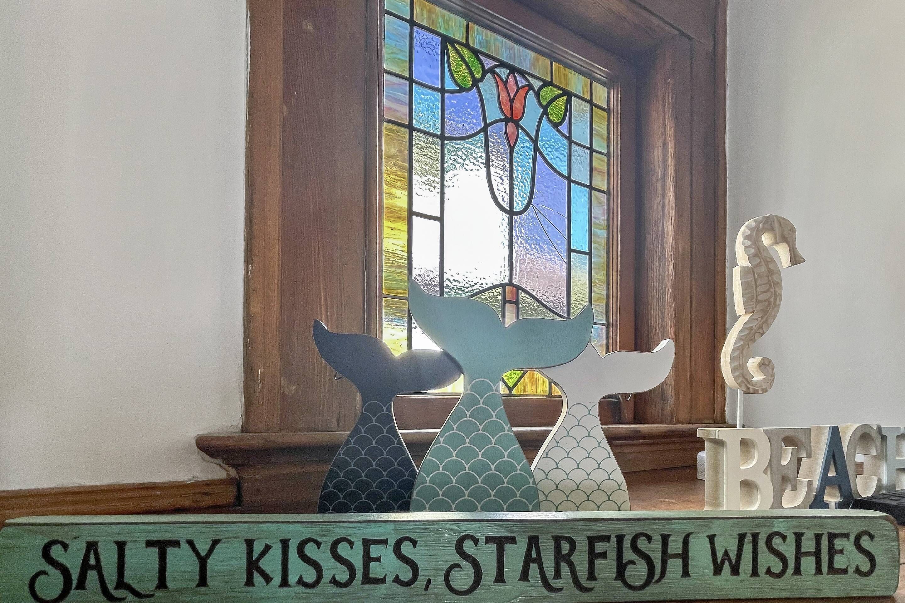 Un letrero de madera con el texto "SALTY KISSES, STARFISH WISHES" frente a una ventana con un diseño de vitral.