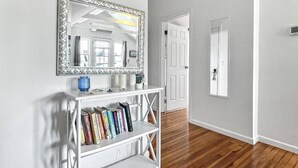 A hallway with a white console table holding books and a blue vase, a large mirror with a decorative frame, and a wooden floor. - Beach Bungalow, walk to beach, private backyard (West Haven)