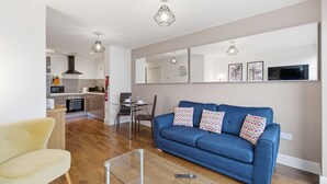 A modern living room with a blue sofa, a glass coffee table, and a kitchen in the background. - Properties Unique | Cherry (Newcastle Upon Tyne)