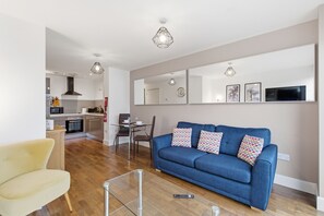 A modern living room with a blue sofa, a glass coffee table, and a kitchen in the background.