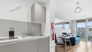 A modern kitchen with a built-in sink, a dining area with a table and chairs, and a blue sofa in a living space with large windows. - Properties Unique | Cherry (Newcastle Upon Tyne)