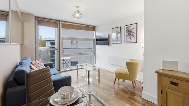 A modern living room with a sofa, dining table, and chairs, large windows, and a view of the city. - Properties Unique | Cherry (Newcastle Upon Tyne)