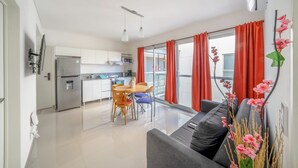 Une cuisine moderne avec des meubles blancs, un réfrigérateur en acier inoxydable et un four à micro-ondes. On y trouve un coin repas avec une table et des chaises, ainsi qu’un balcon agrémenté de rideaux rouges.