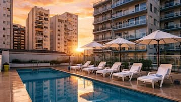 Una piscina en la azotea con sillones y sombrillas, rodeada de edificios altos.