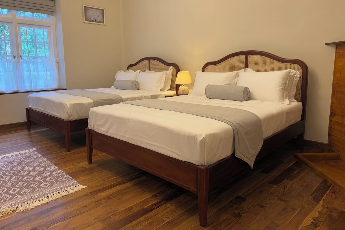 Une chambre d’hôtel avec deux lits en bois, une petite table de chevet avec une lampe, une fenêtre avec des rideaux et un tapis à motifs sur le sol.