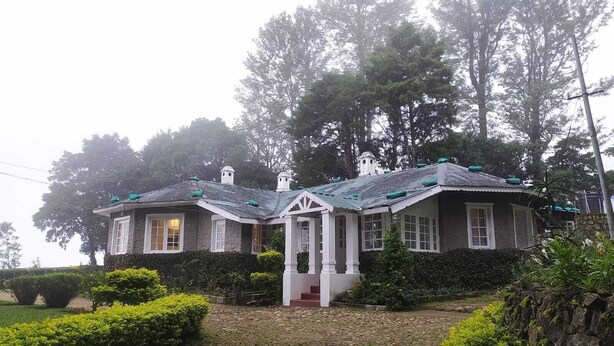 A traditional stone house with a gabled roof, a central entrance, and a surrounding garden. - The Gundumalai Bungalow  (Suryanelli)