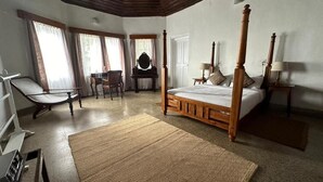 A spacious bedroom with a four-poster bed, a sitting area with a chair and table, and a large window with curtains. - The Gundumalai Bungalow  (Suryanelli)