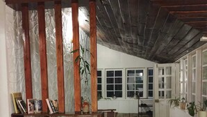 A spacious room with a high ceiling, wooden beams, and a large window. There is a wooden bench with a vase of flowers and a bookshelf. - The Gundumalai Bungalow  (Suryanelli)
