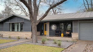 A modern house with a garage, a large tree in the front yard, and a small patio with chairs and a table.