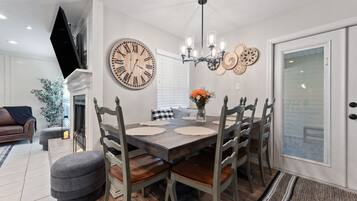 A dining area with a wooden table, six chairs, a large wall clock, and a chandelier.