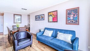 A living room with a blue sofa, a black leather chair, and a dining area with a wooden table and chairs. - Increíble depto a pasos de Av. del Mar (La Serena)
