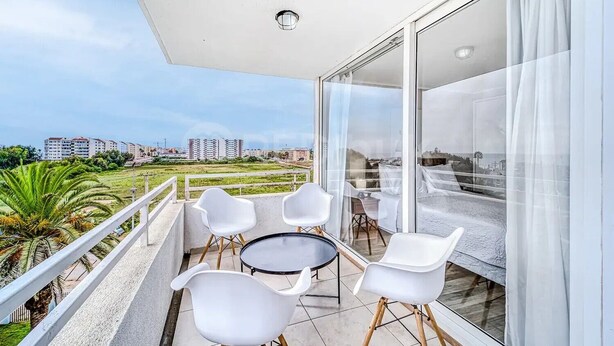 A balcony with a glass door, white chairs, a round table, and a view of buildings and greenery. - Increíble depto a pasos de Av. del Mar (La Serena)