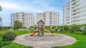 A playground with a wooden slide and two colorful slides, surrounded by a circular concrete area, with apartment buildings in the background. - Increíble depto a pasos de Av. del Mar (La Serena)